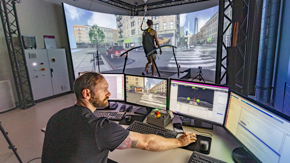 Das Ganganalyse-Labor (GRAIL) der TU Chemnitz ermöglicht Grundlagen- und Anwendungsforschung innerhalb einer interaktiven Echtzeit-Virtual-Reality-Umgebung. Die virtuelle Welt wird auf einen 240 Grad „curved Screen“ projiziert. Damit die Probandin während der Ganganalyse auf dem Laufband nicht stürzt, ist sie mit einem Gurt gesichert. 